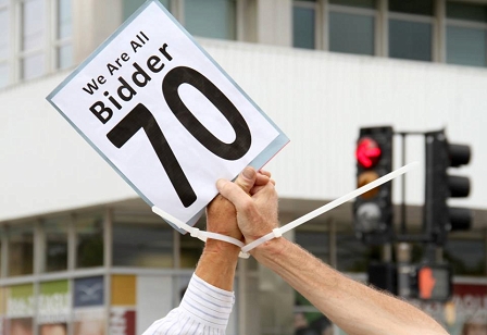 Zwei mit Kabelbinder gefesselte Hände halten ein Schild hoch mit der Aufschrift "We are all Bidder 70"